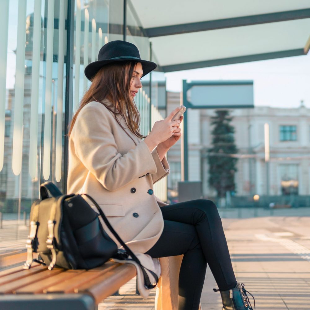 Tourist girl sits at public transport stop and looks in smartphone. Girl waiting for the bus